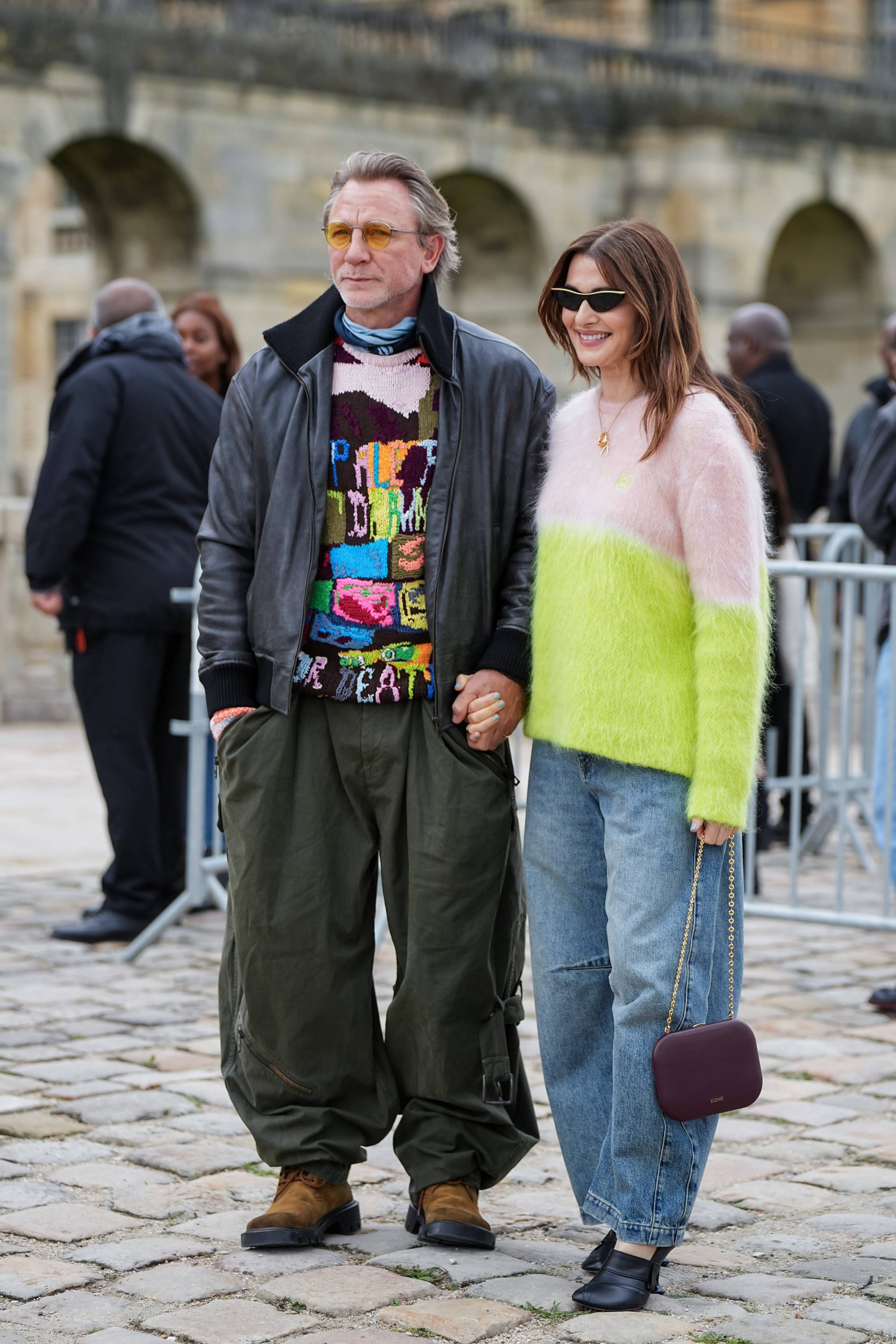 Daniel Craig and Rachel Weisz at the Loewe fashion show. He is wearing a black bomber jacket, unusual patterned jumper and khaki trousers. She is wearing a pink and yellow jumper and a baggy blue jeans